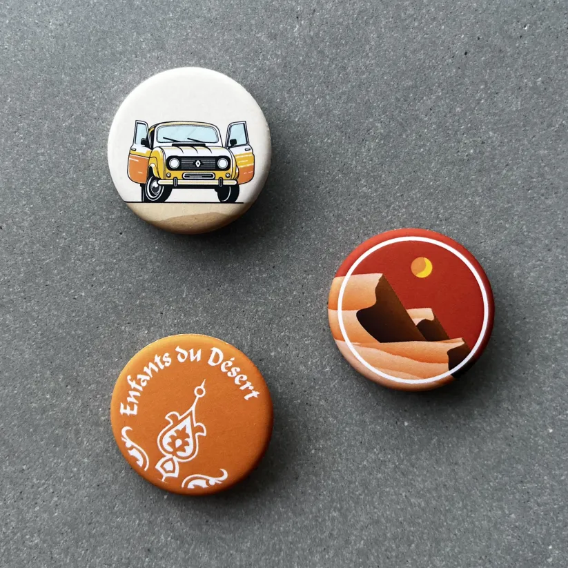 Badges illustrés Enfants du Désert avec voiture, paysage de dunes et logo de l’association.